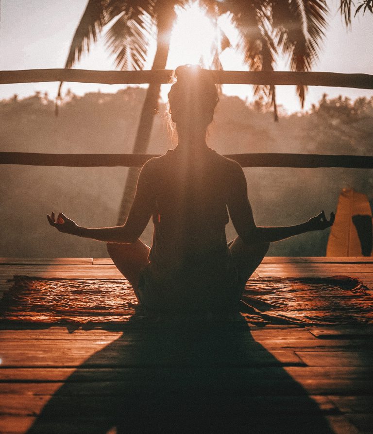 A woman in a yoga pose sits silhouetted against a sunset beach palm tree background.
