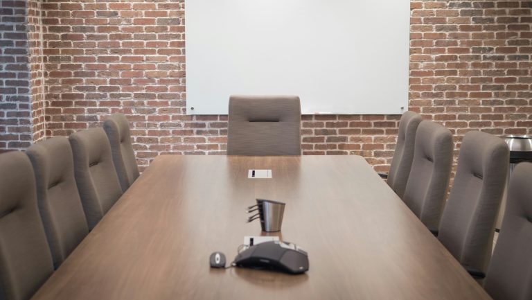 An empty meeting desk with chairs around it in a brick walled room with a white board on one side.