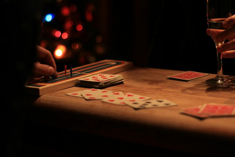 A dark and moody playing card game of cribbage, with soft lighting on the wooden table and a hand holding a glass of wine.