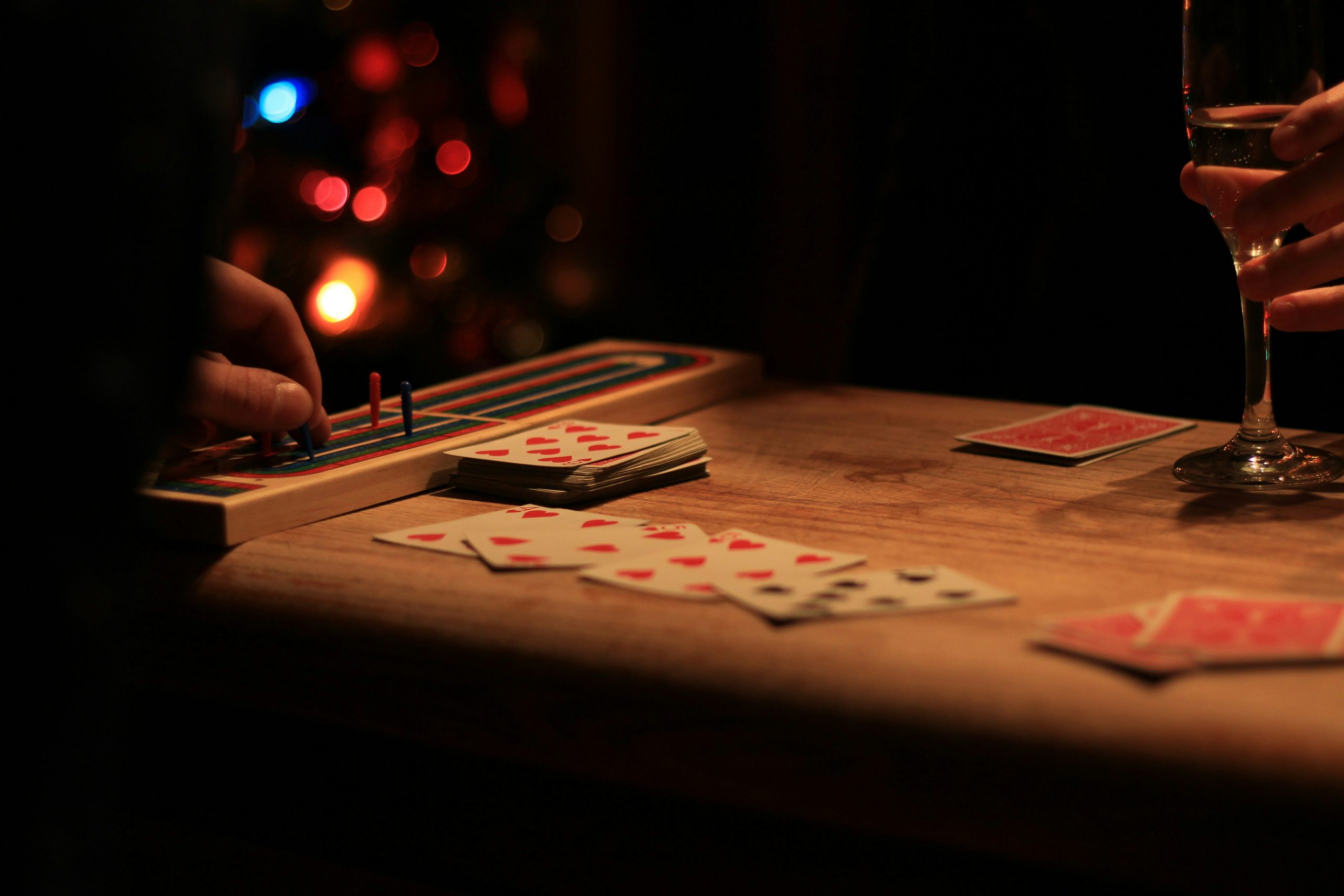 A dark and moody playing card game of cribbage, with soft lighting on the wooden table and a hand holding a glass of wine.