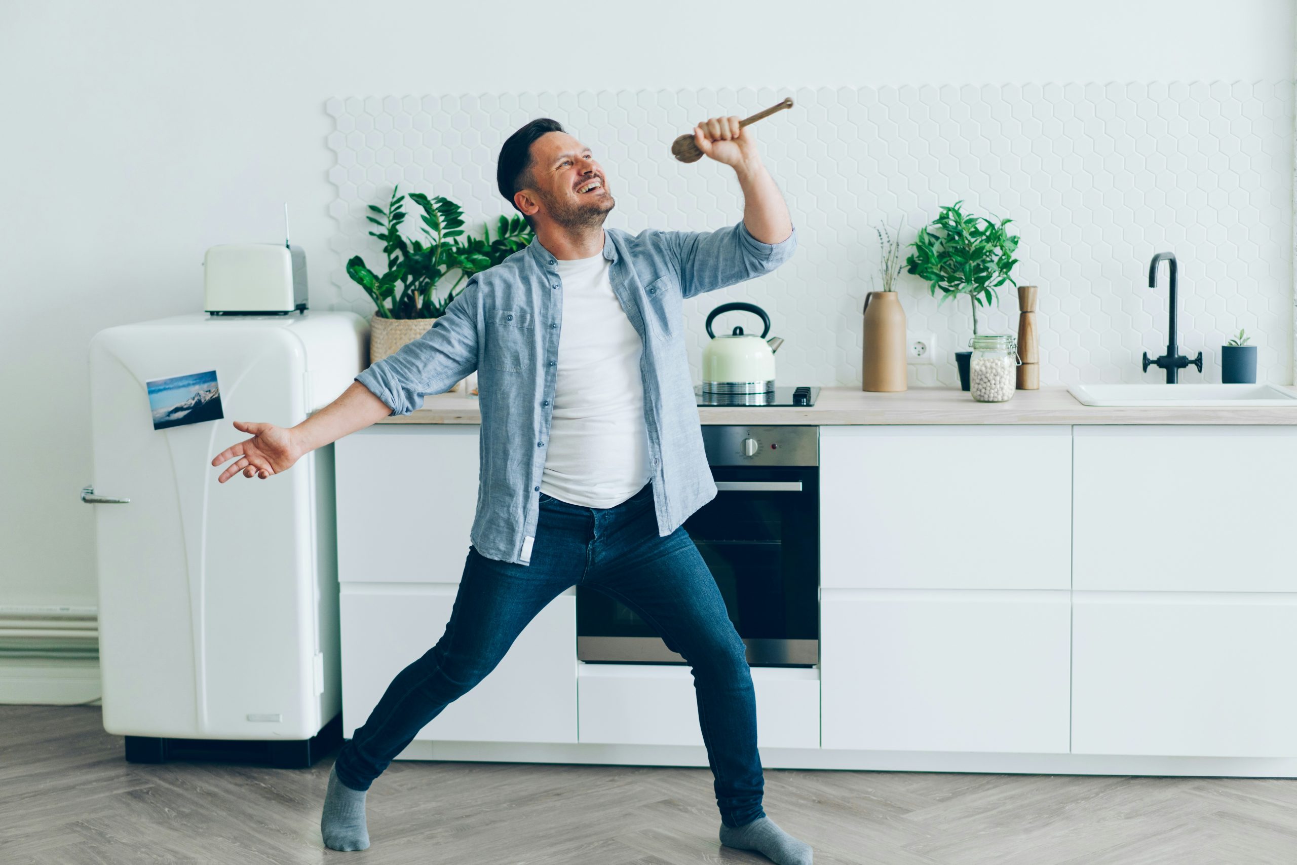 A man in a modern white kitchen posing with a microphone as if he is singing very enthusiastically.